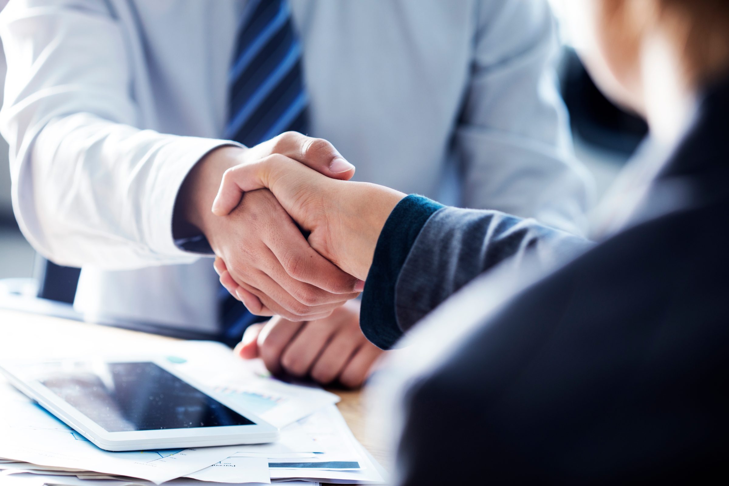 Two business professionals shaking hands over documents and a tablet at a desk