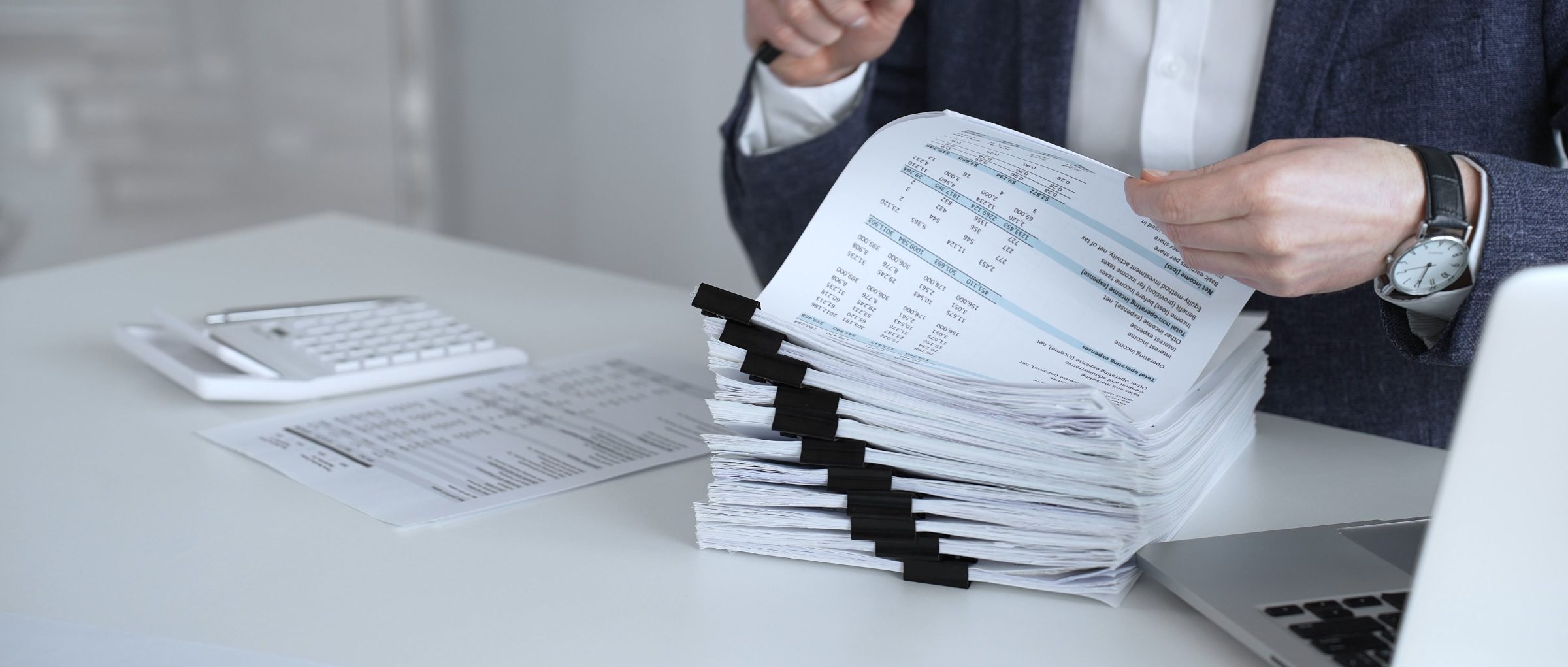 Businessperson holding stack of financial documents with data tables at desk with calculator and laptop
